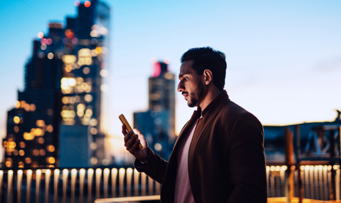Businessman Using Mobile Phone At Rooftop In Business Building Against Illuminated Financial Buildings And London Cityscape At Night
